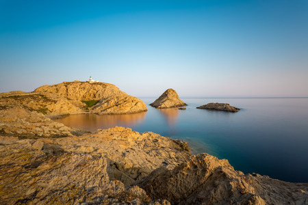 Red rocks and lighthouse at Ile Rousse in Balagne region of Corsica above a mirror like and calm...