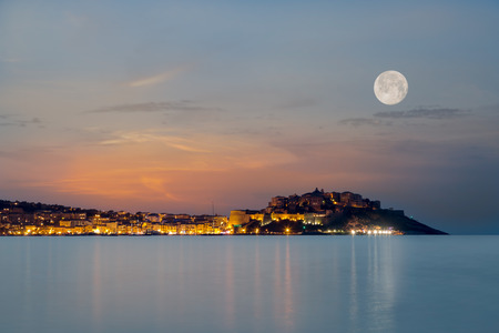 Full moon over the citadel of Calvi in the Balagne region of Corsica at dusk with reflections of light across a calm mediterranean seaの写真素材