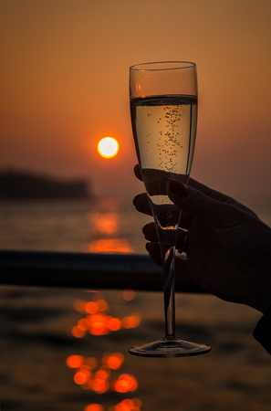 Orange glowing sun setting behind a glass of champagne held up to the light with bubbles rising through reflected lightの写真素材