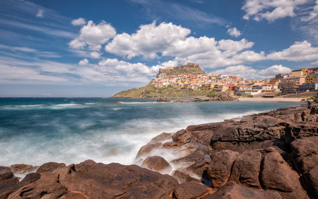Slow shutter image of waves crashing onto rocks at Castelsardo on the coast of Sardinia in Italy with rocks in the foreground and blue skies and fluffy cloudsの写真素材