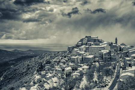 Black & White image of the snow covered mountain village of Speloncato in the Balagne region of Corsica with the Mediterranean behind and dark clouds aboveの写真素材