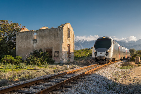 Train passing a derelict station at Lumio in the Balagne region of Corsica with snow covered mountains in the distance under a depp blue skyの写真素材