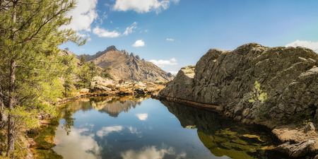 Panoramic view of small lake at Paglia Orba surrounded by rocks, pine trees and mountains near the GR20 hiking trail in central Corsicaの写真素材