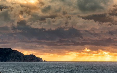 Evening sun rays bursting through dark stormy clouds onto the Mediterranean sea behind the Revellata lighthouse near Calvi in the Balagne region of Corsicaの写真素材