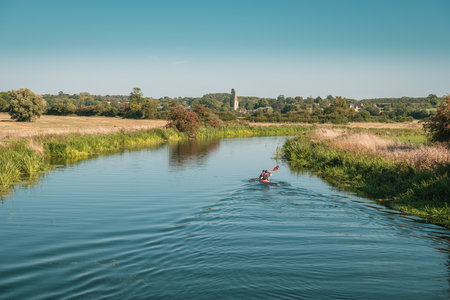 WOODFORD, NORTHAMPTONSHIRE,ENGLAND - 2nd SEPTEMBER 2018. Two canoeists in a double kayak paddle along a calm River Nene near Woodford Lock in Northamptonshire with Denford church in the distance on a bright sunny dayのeditorial素材