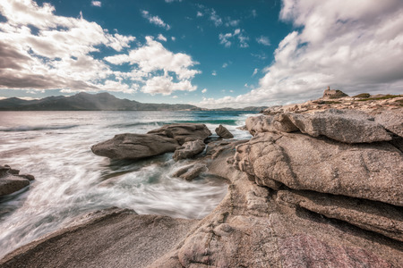 Dramatic long exposure image of waves washing onto rocks on the coast by the Genoese tower at Punta Caldanu near Lumio in the Balagne region of Corsica with Calvi in the distanceの写真素材