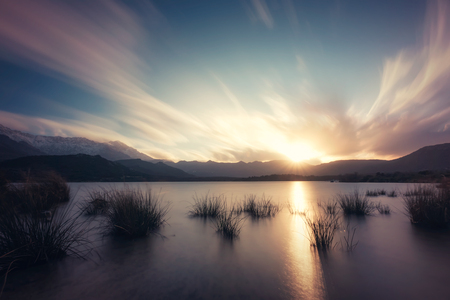 Long exposure image of sun setting behind clouds over Lac de Codole in the Regino valley in the Balagne region of Corsica with reeds in the foreground and snow capped mountains in the distanceの写真素材