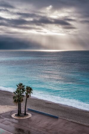 Three palm trees stand next to an empty row of chairs on the sea front on the Promenade des Anglais in Nice against the turquoise Mediterranean sea with sun rays filtering through dark storm clouds in the distanceの写真素材