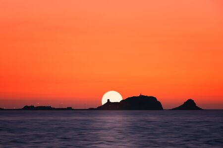 Sun setting behind a silhoutted  "La Pietra", the red rock at Ile Rousse in the Balagne region of Corsicaの写真素材