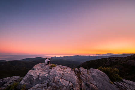 A man and his border collie dog enjoy the view of the coastline and the Mediterranean sea at sunrise from a rocky mountain outcrop at the Col de San Colombano in Corsicaの写真素材