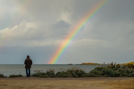 Man in hooded coat looking out across the Mediterranean sea towards a colourful rainbowの写真素材