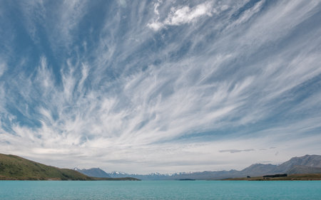 Wispy clouds and blue skies over the turquoise blue water of Lake Tekapo in New Zealand with snow capped mountains in the distanceの写真素材