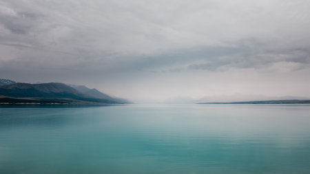 Clouds and mist hang over over the turquoise blue water of Lake Tekapo in New Zealand with snow capped mountains in the distanceの写真素材