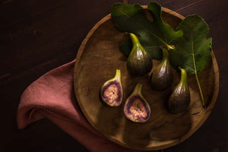 Juicy fresh whole fig fruits and one cut in half on round olive wood platter on dark wood background...