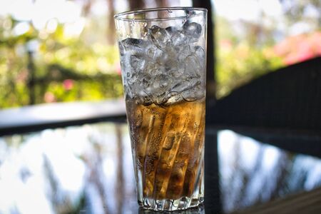 This unique photo shows an almost full glass of cola with ice. The drink is on a glass table in the garden of a restaurant in Hua Hin in Thailandの写真素材