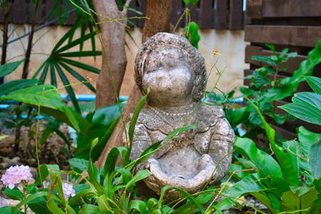 This unique photo shows an old weathered stone statue which is supposed to represent a fat woman. There are many leaves and a flower around the figureの写真素材