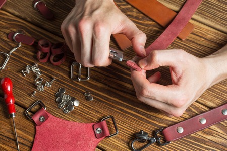 The master holds in his hands a knife and a piece of leather. On brown wooden table scattered with...
