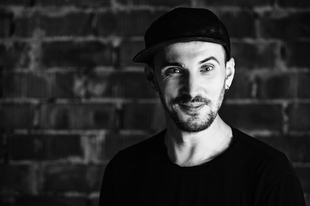 Young man wearing a black t-shirt and black cap a brick wall. Black and white photography....