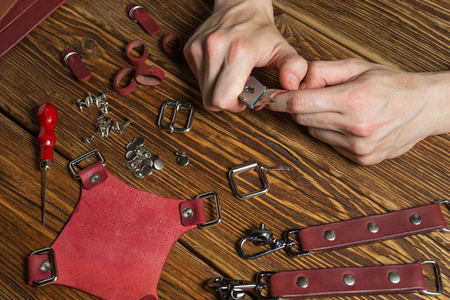 The master holds in his hands a knife and a piece of leather. On brown wooden table scattered with...