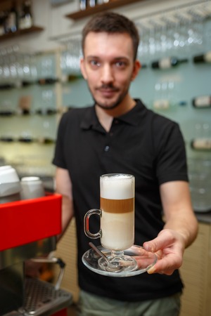 Men barista hold glass of latte serving a client. Focus on beverage. Shallow depth of field. Vertically framed shot.の写真素材