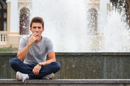 The guy smokes a cigarette sitting crossed legs about fountain. Horizontally framed shot.の写真素材
