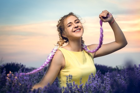 Portrait of a beautiful girl with long braids in a lavender field. Holds pigtails in his hands. Sunset in the background. Horizontally framed shot.の写真素材