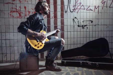 Street musician sitting on the amplifier and playing guitar in the underpass. Vagrant lifestyle. Playing in the underpass to make a living. Unemployed musician. Future rock star. Horizontally framed shot.の写真素材