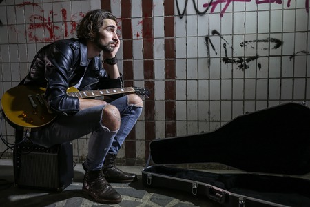 Street musician sitting on the amplifier and playing guitar in the underpass. Vagrant lifestyle. Playing in the underpass to make a living. Unemployed musician. Future rock star. Horizontally framed shot.の写真素材