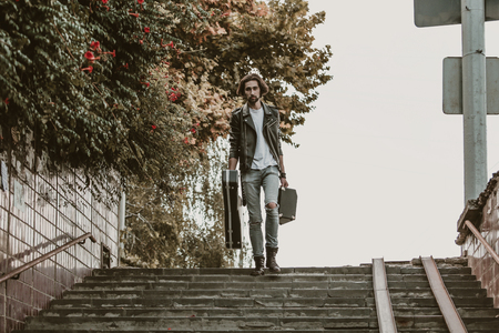 Street musician holding a case with a guitar and amplifier. Goes down the stairs to the underground passage. Vagrant lifestyle. Playing to make money a living. Unemployed musician. Future rock star. Horizontally framed shot.の写真素材