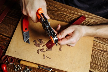 Master holding a hole punch and a piece of leather. On brown wooden table scattered with tools and...