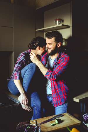 Beautiful young couple in kitchen at home while cooking healthy food. Husband cuts salad and feeds his wife cucumber. They both laugh.の写真素材