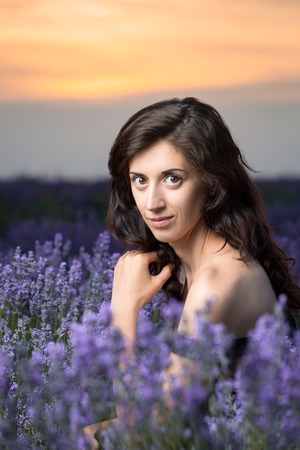 Portrait of a beautiful brunette in a black dress in a lavender field. Sunset in the background. Vertically framed shot.の写真素材