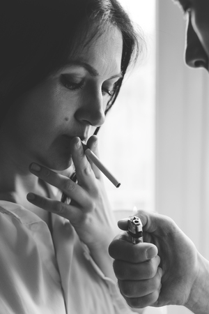 Young man light in a cigarette to a young woman. Black and white photo. Vertically framed shot.の写真素材