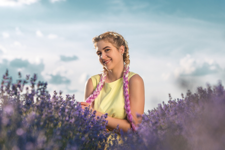 A young, pretty blonde with long pink braids stands in a lavender field. Horizontally framed shot.の写真素材