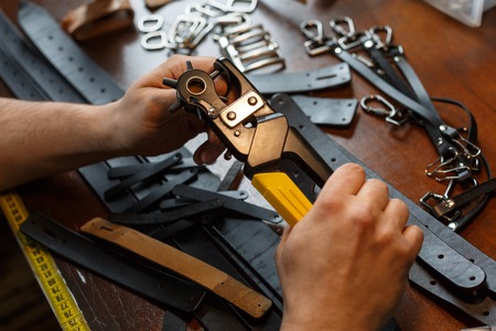 Master holding a hole punch and a piece of leather. On brown wooden table scattered with tools and accessories. Horizontally framed shot.の写真素材