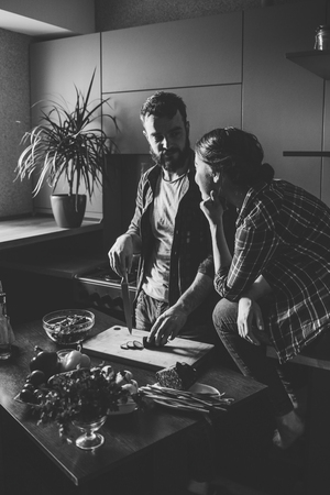 Beautiful young couple in kitchen at home while cooking healthy food. Man is cuts salad. Woman sits on table and he looks at her husband.の写真素材