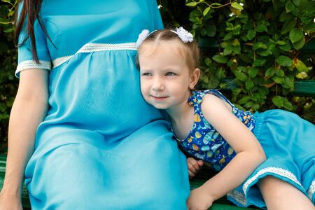 Little girl sits on a bench and leaned against her mother's stomach. Woman pregnant. Horizontally framed shot.の写真素材