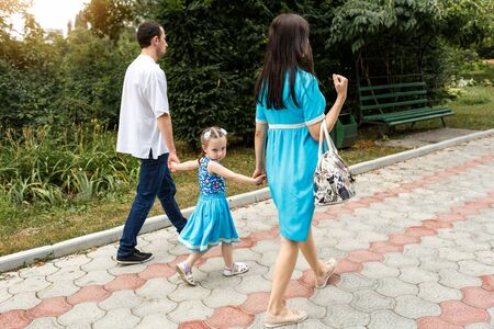 Back view on happy family walking on path holding hands and spending time in beautiful green park. Horizontally framed shot.の写真素材