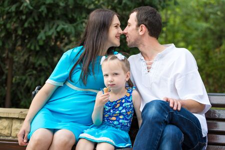 Family sitting on a bench in park. Little girl eating a waffle cup. Woman pregnant. Happy family life concept. Horizontally framed shot.の写真素材