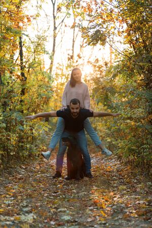 Young beautiful couple and their labrador in the Park. A man holds a woman on his back, and they spread their arms. Rays of setting sun break through the branches of trees. Vertically framed shot.の写真素材