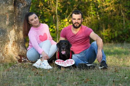 Young couple sitting on the grass in the Park. Between them is a black labrador with a sign in the...