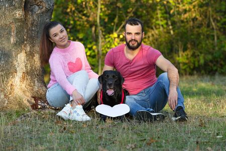 Young couple sitting on the grass in the Park. Between them is a black labrador with a white sign in the shape of a heart. The concept of a happy family life in love. Horizontally framed shot.の写真素材