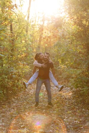 Young beautiful couple and their labrador in the Park. A man holds a woman on his back and she hugs him. Rays of setting sun break through the branches of trees. Vertically framed shot.の写真素材