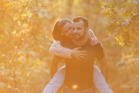 Young beautiful couple and their labrador in the Park. A man holds a woman on his back and she hugs him. Warm autumn evening in the forest. Horizontally framed shot.の写真素材