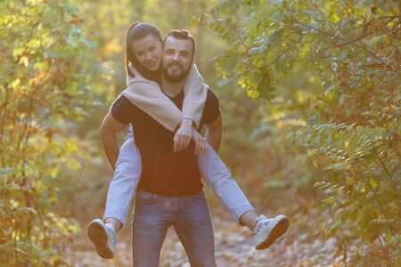 Young beautiful couple and their labrador in the Park. A man holds a woman on his back and she hugs him. Warm autumn evening in the forest. Horizontally framed shot.の写真素材