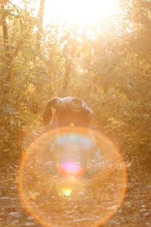 Young beautiful couple and their labrador in the Park. A man holds a woman on his back and she hugs him. Rays of setting sun break through the branches of trees. Vertically framed shot.の写真素材