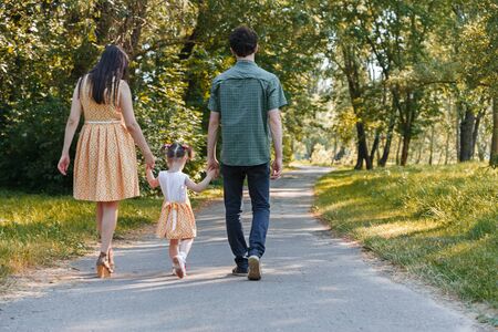 Back view on happy family walking on path holding hands and spending time in beautiful green park. Horizontally framed shot.の写真素材