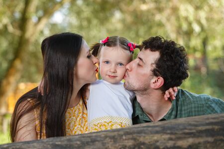 Family portrait of parents and daughter. A man and a woman kiss the girl on the cheeks. Nature in the background. Horizontally framed shot.の写真素材