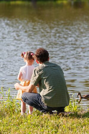 Father and daughter looking at lake. A wild duck swims through the water. Back view. Vertically framed shot.の写真素材