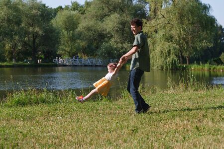 Dad playing with his baby daughter in a yellow dress. He turns her around himself at arm's length. In the background a lake and a bridge. Horizontally framed shot.の写真素材